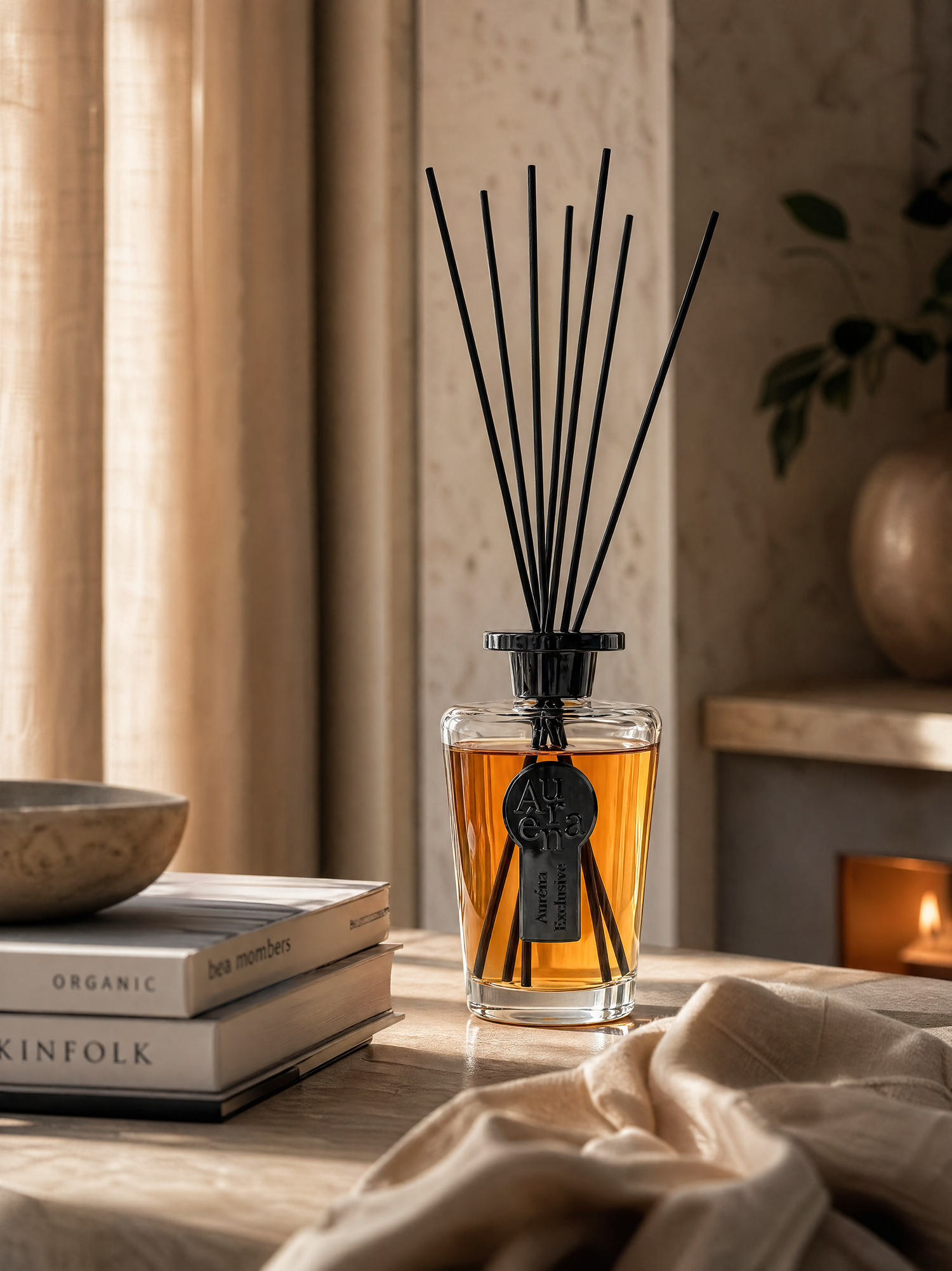 Eleganter Auréna Raumduft-Diffuser mit schwarzem Label und langen Duftstäbchen steht auf einer hellen Holzoberfläche in einem warm beleuchteten, stilvollen Interieur. Neben dem Diffuser liegen dekorative Bücher und eine Schale, im Hintergrund sorgt eine brennende Kerze für eine ruhige, luxuriöse Atmosphäre mit weichen Lichtreflexen und natürlichen Texturen.